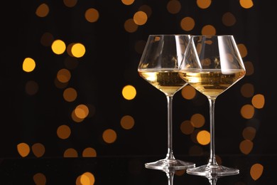 Photo of Tasty white wine in glasses on mirror surface against dark background with blurred lights, space for text. Bokeh effect