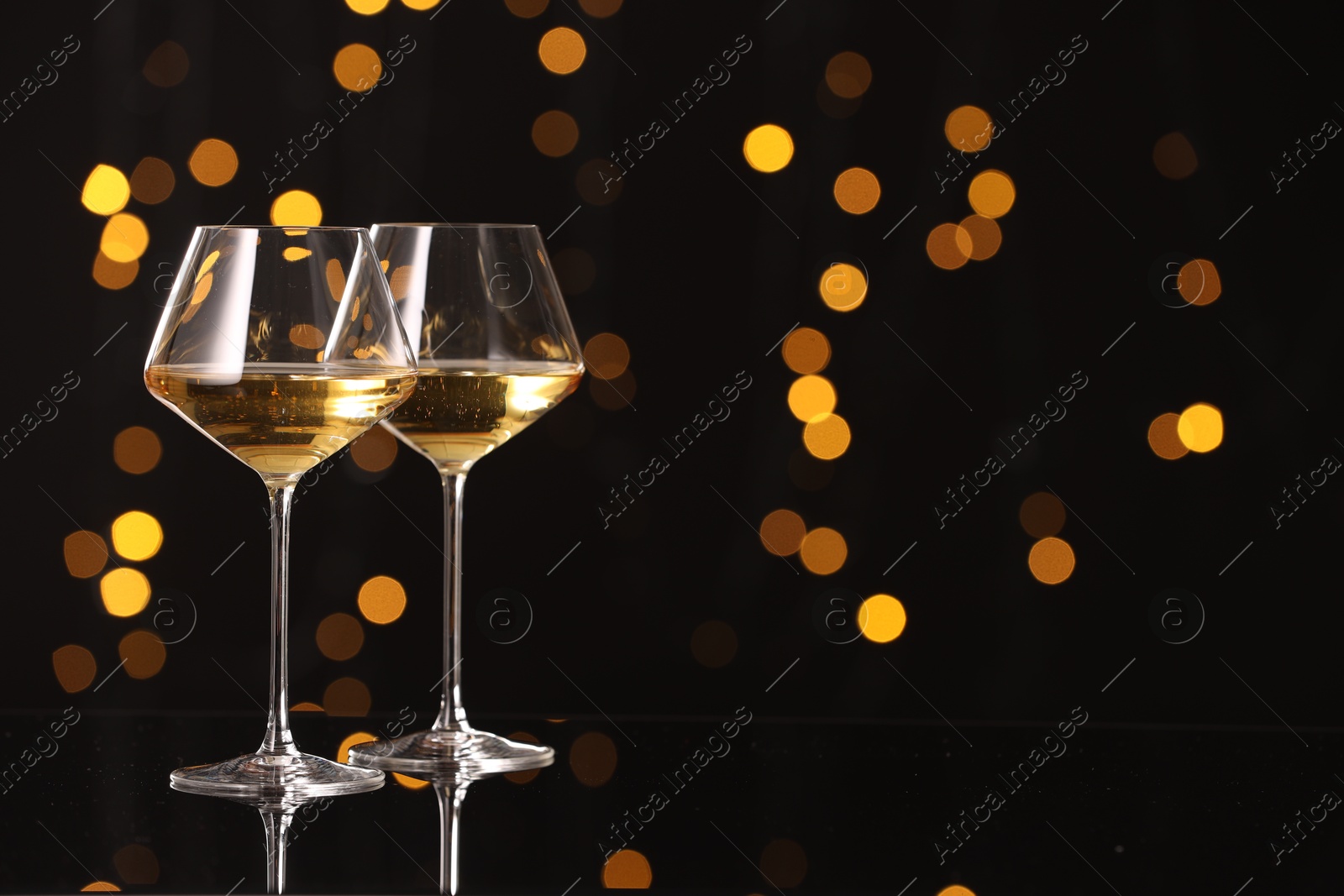 Photo of Tasty white wine in glasses on mirror surface against dark background with blurred lights, space for text. Bokeh effect