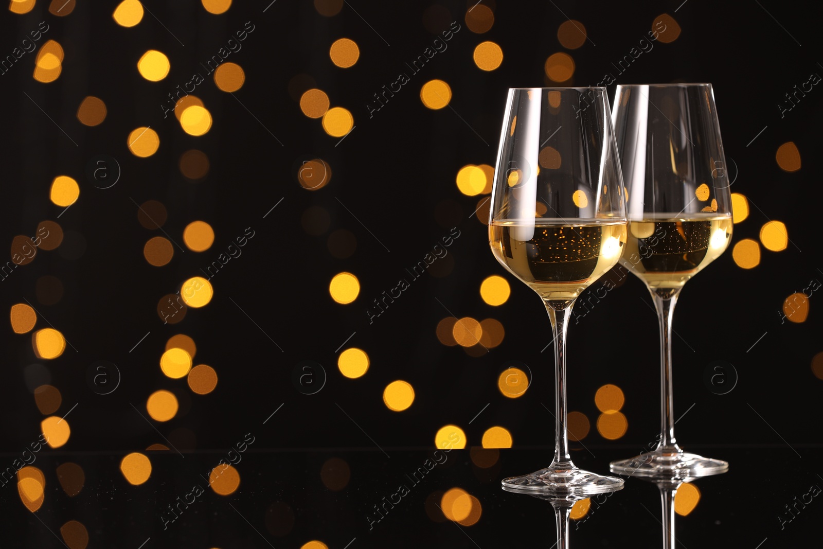 Photo of Tasty white wine in glasses on mirror surface against dark background with blurred lights, space for text. Bokeh effect