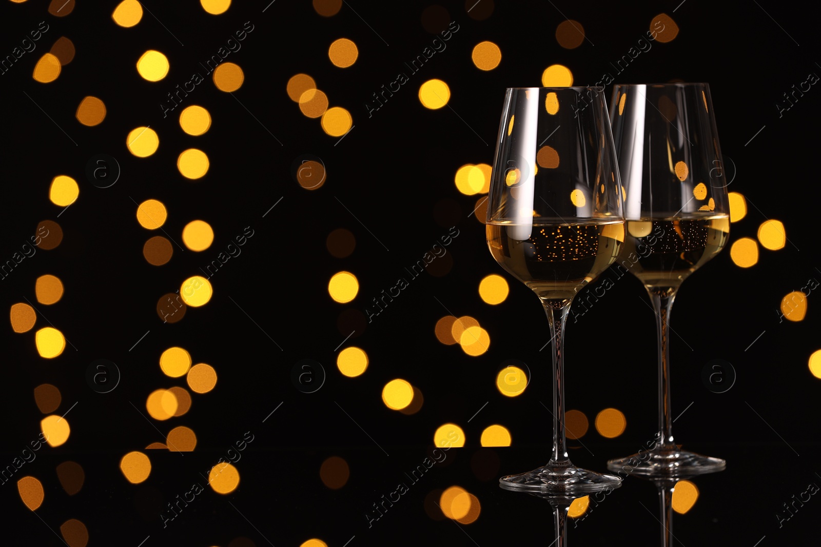 Photo of Tasty white wine in glasses on mirror surface against dark background with blurred lights, space for text. Bokeh effect