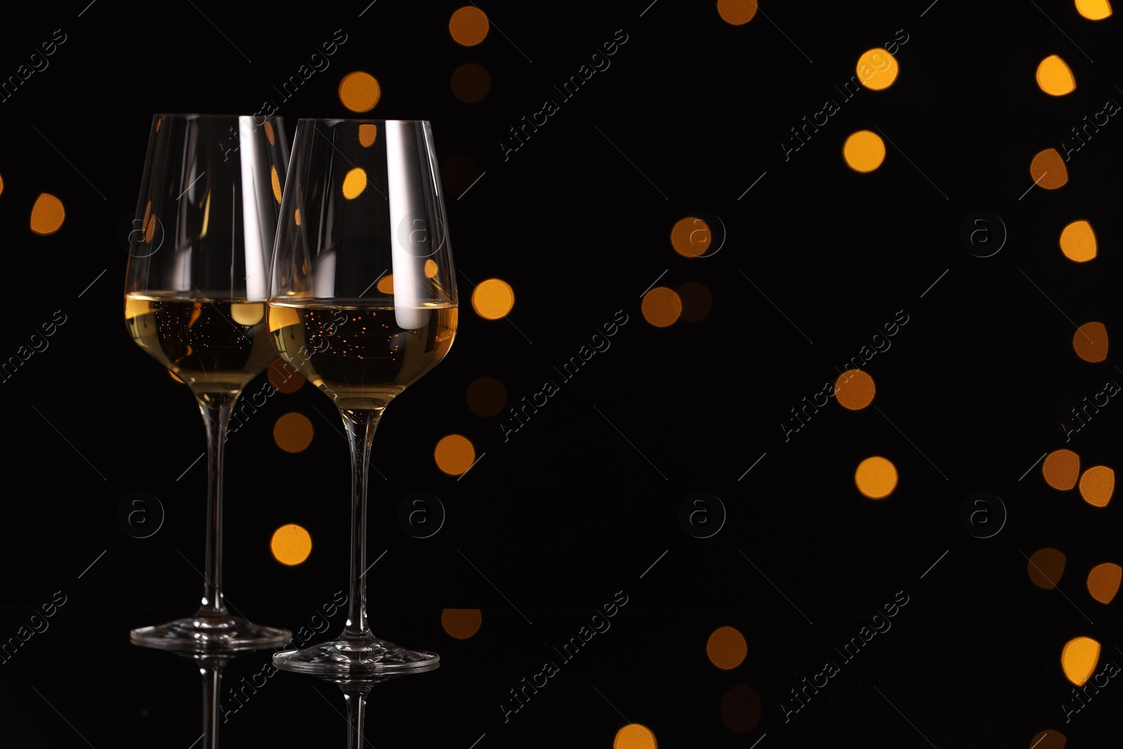 Photo of Tasty white wine in glasses on mirror surface against dark background with blurred lights, space for text. Bokeh effect