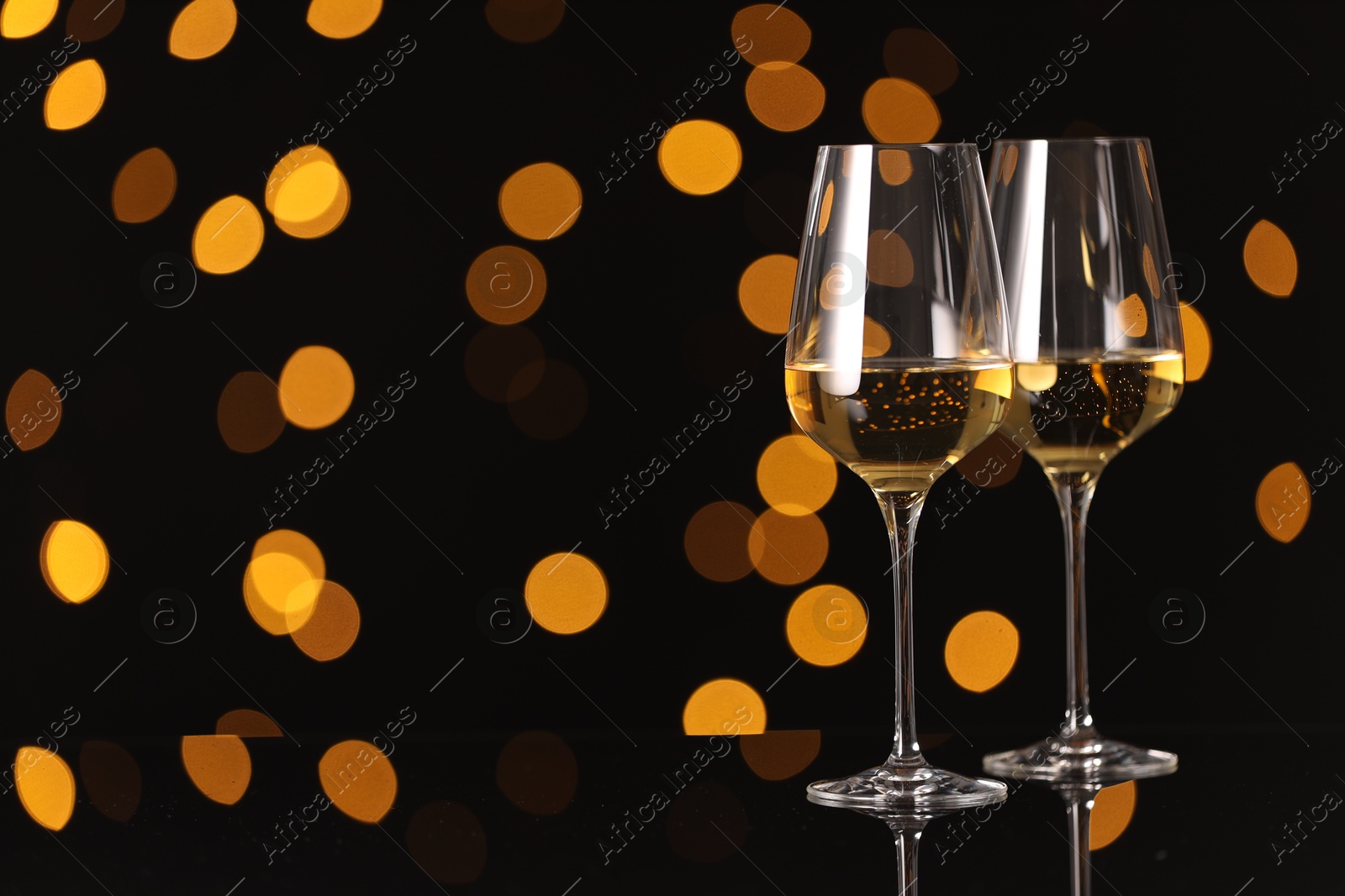 Photo of Tasty white wine in glasses on mirror surface against dark background with blurred lights, space for text. Bokeh effect