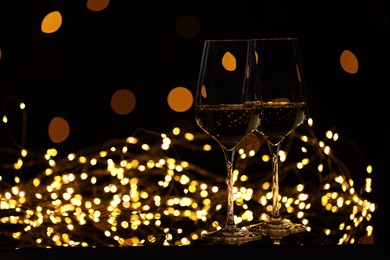 Photo of Tasty white wine in glasses and Christmas lights against black background, bokeh effect