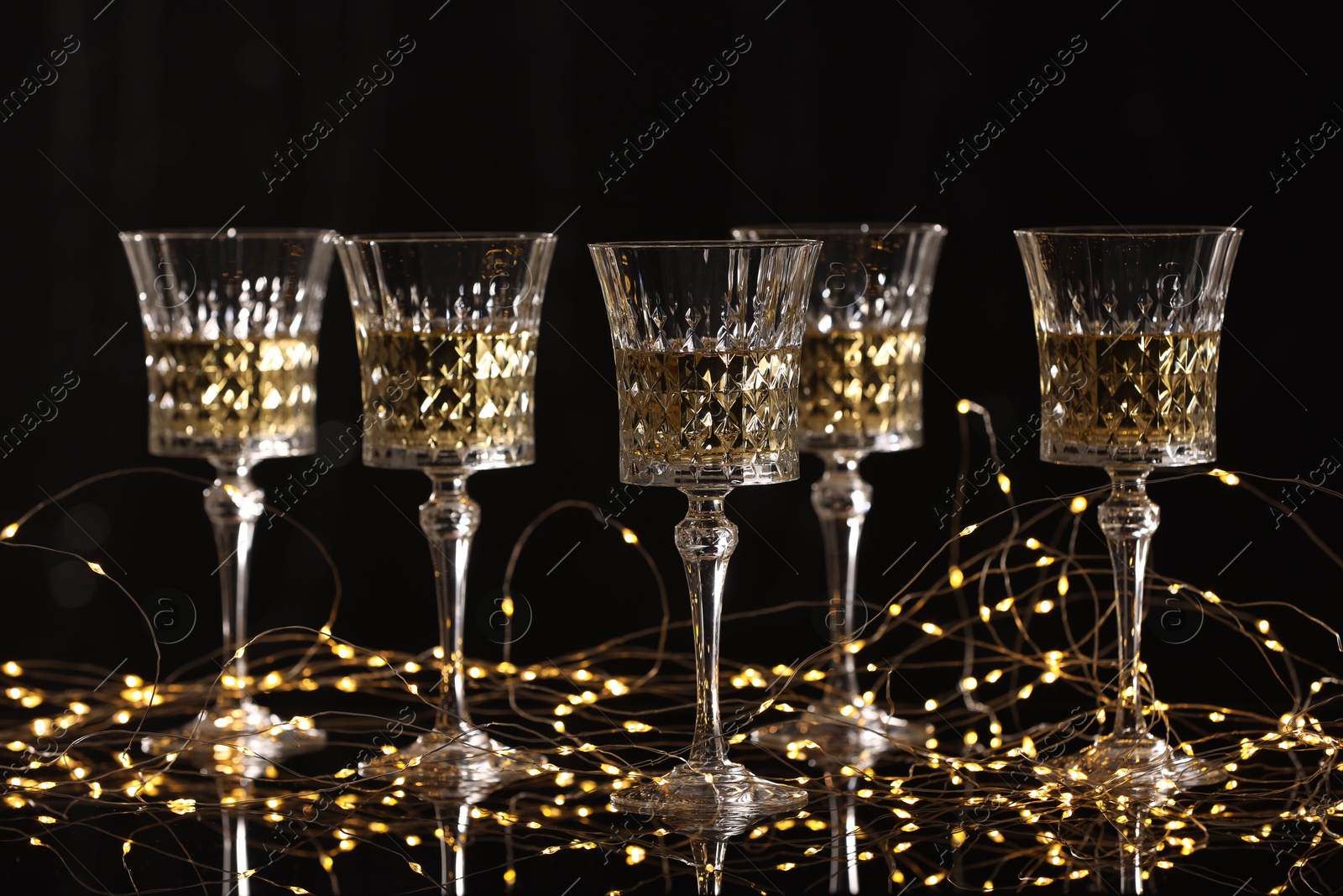 Photo of Tasty white wine in glasses and Christmas lights against black background