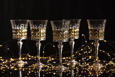 Photo of Tasty white wine in glasses and Christmas lights against black background