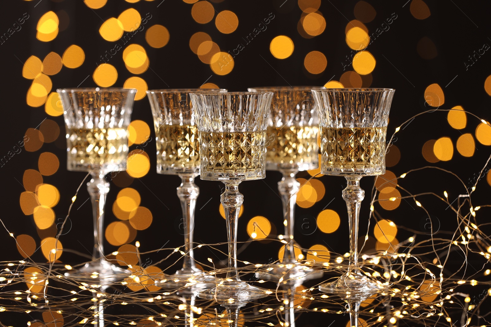 Photo of Tasty white wine in glasses and Christmas lights against dark background, bokeh effect