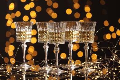 Photo of Tasty white wine in glasses and Christmas lights against dark background, bokeh effect