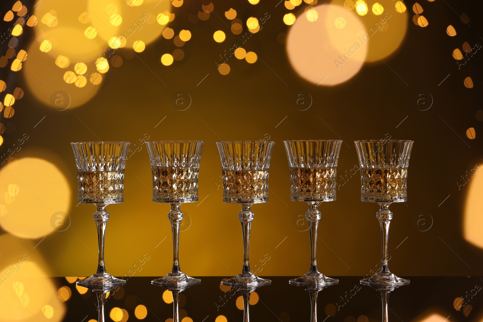 Photo of Tasty white wine in glasses on mirror surface against golden background with blurred lights. Bokeh effect