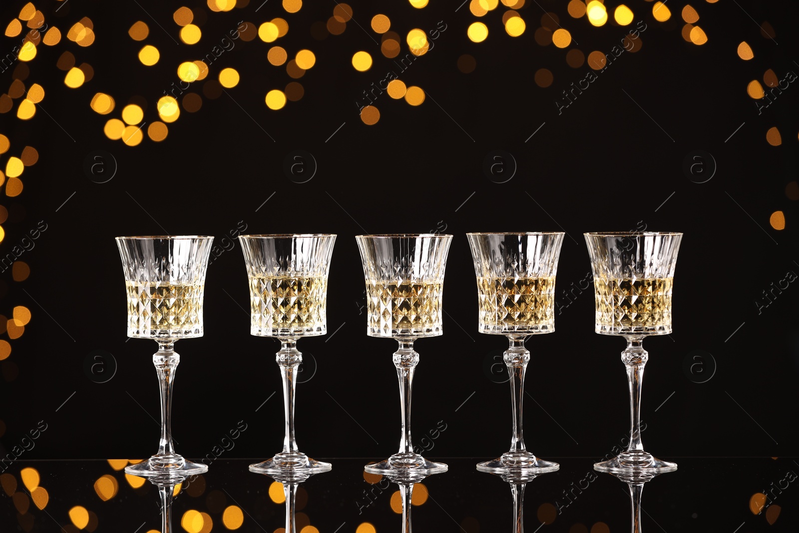 Photo of Tasty white wine in glasses on mirror surface against dark background with blurred lights. Bokeh effect