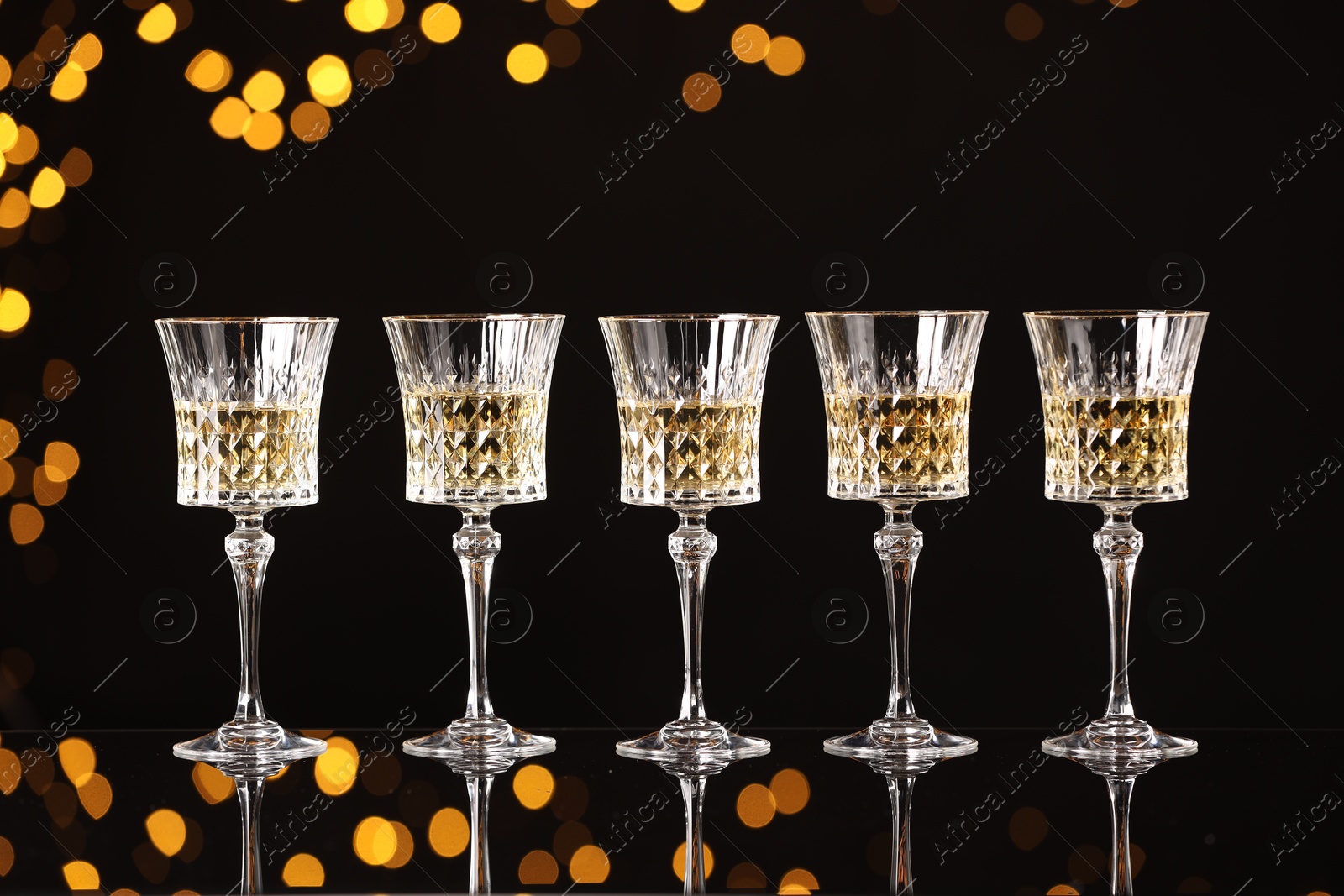 Photo of Tasty white wine in glasses on mirror surface against dark background with blurred lights. Bokeh effect