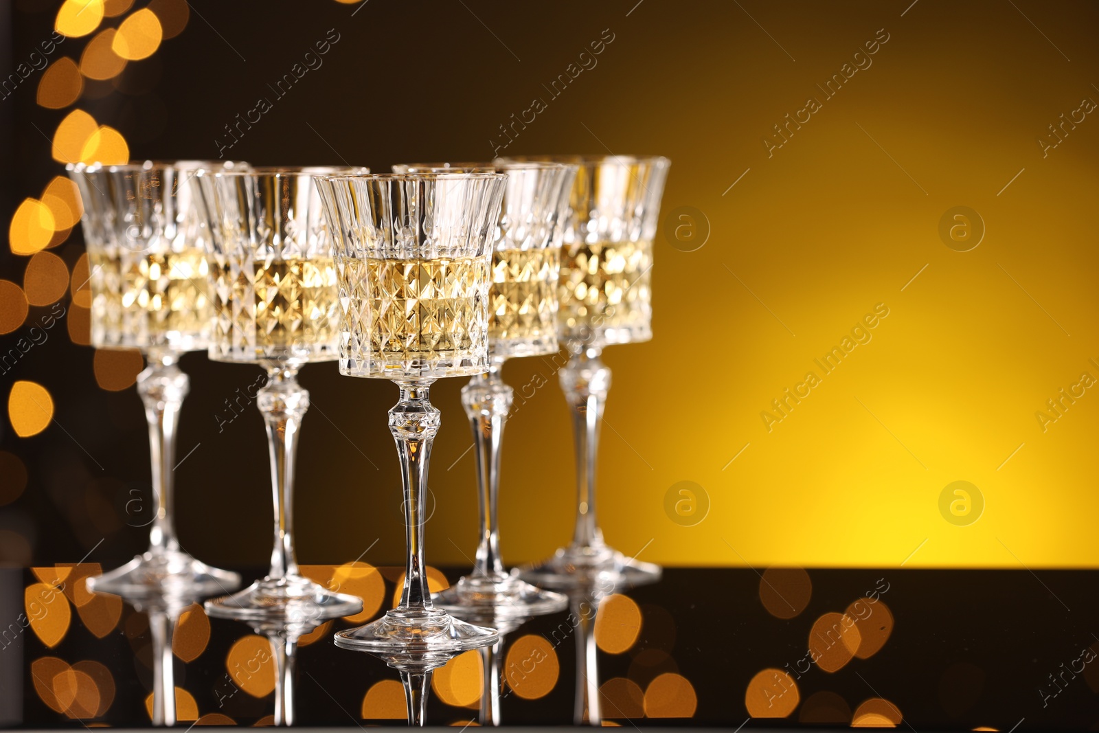 Photo of Tasty white wine in glasses on mirror surface against golden background with blurred lights, space for text. Bokeh effect