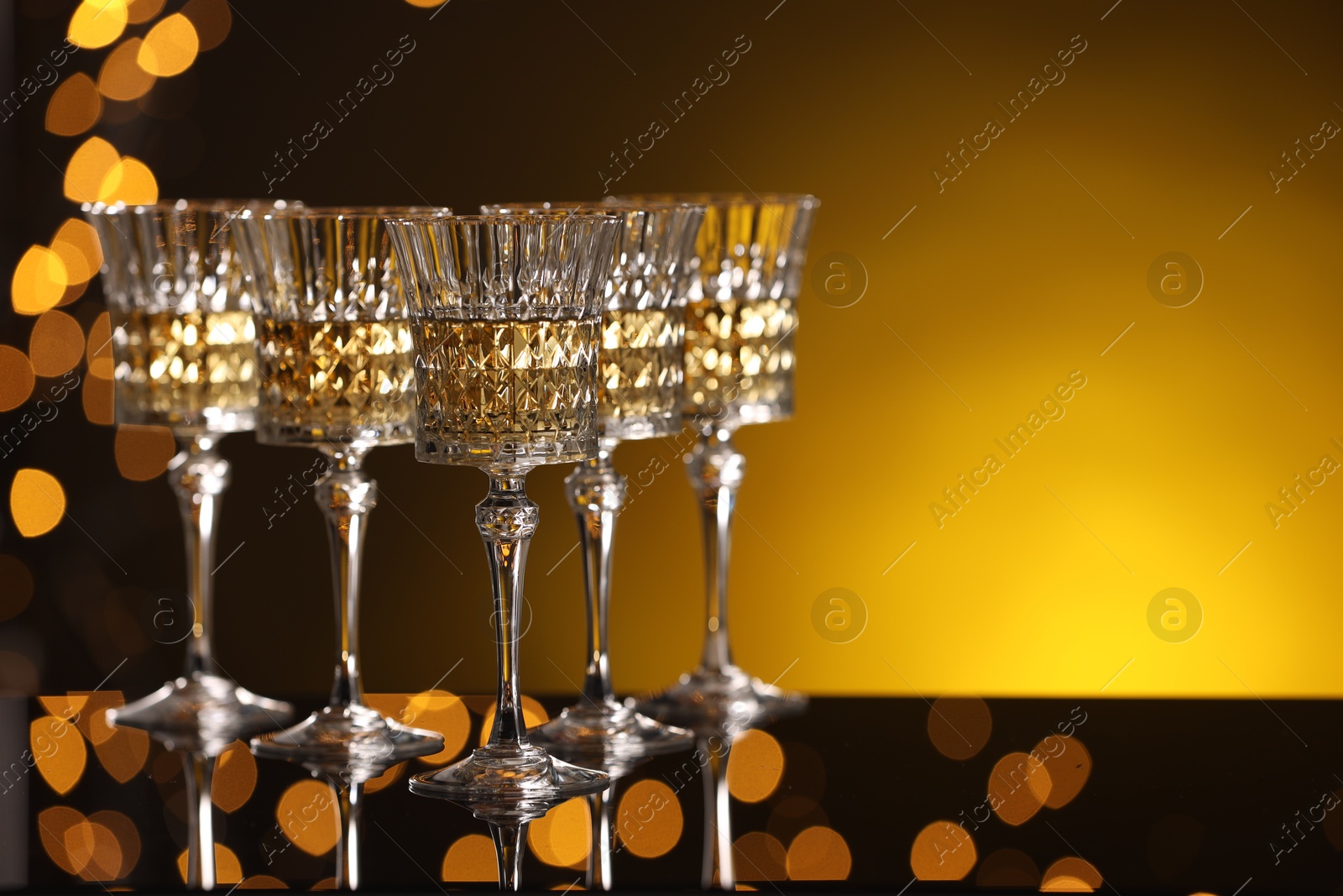 Photo of Tasty white wine in glasses on mirror surface against golden background with blurred lights, space for text. Bokeh effect