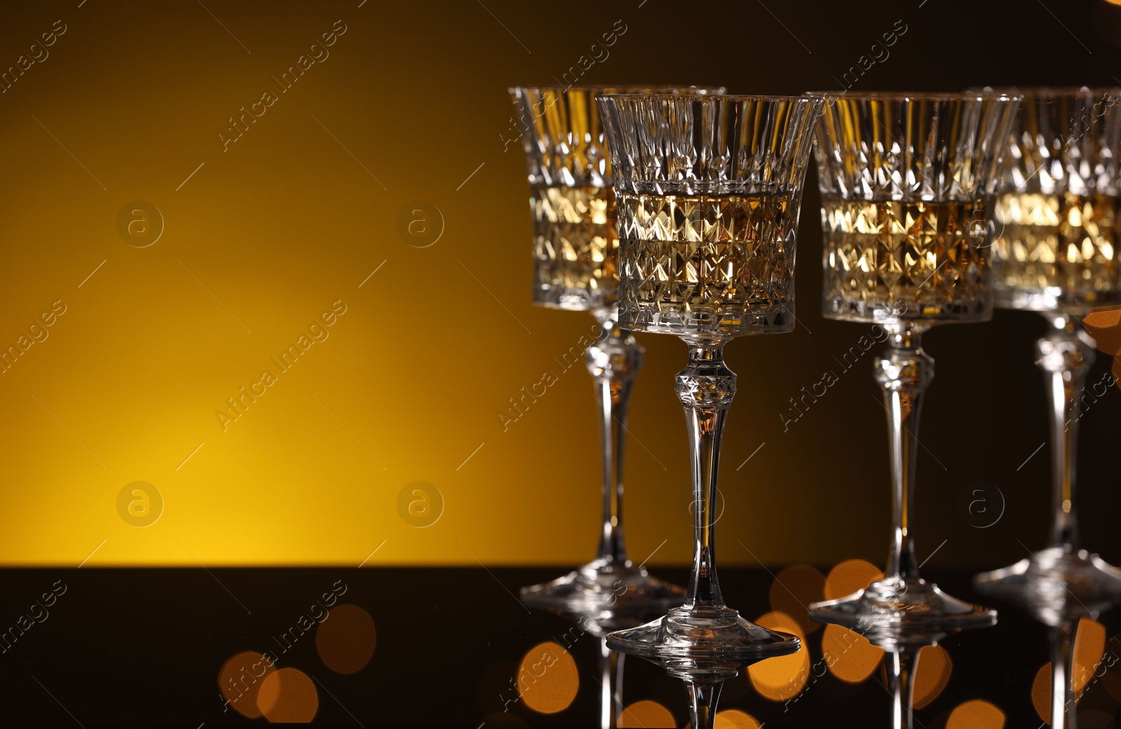 Photo of Tasty white wine in glasses on mirror surface against golden background with blurred lights, space for text. Bokeh effect