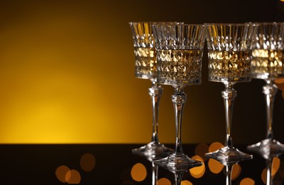 Photo of Tasty white wine in glasses on mirror surface against golden background with blurred lights, space for text. Bokeh effect