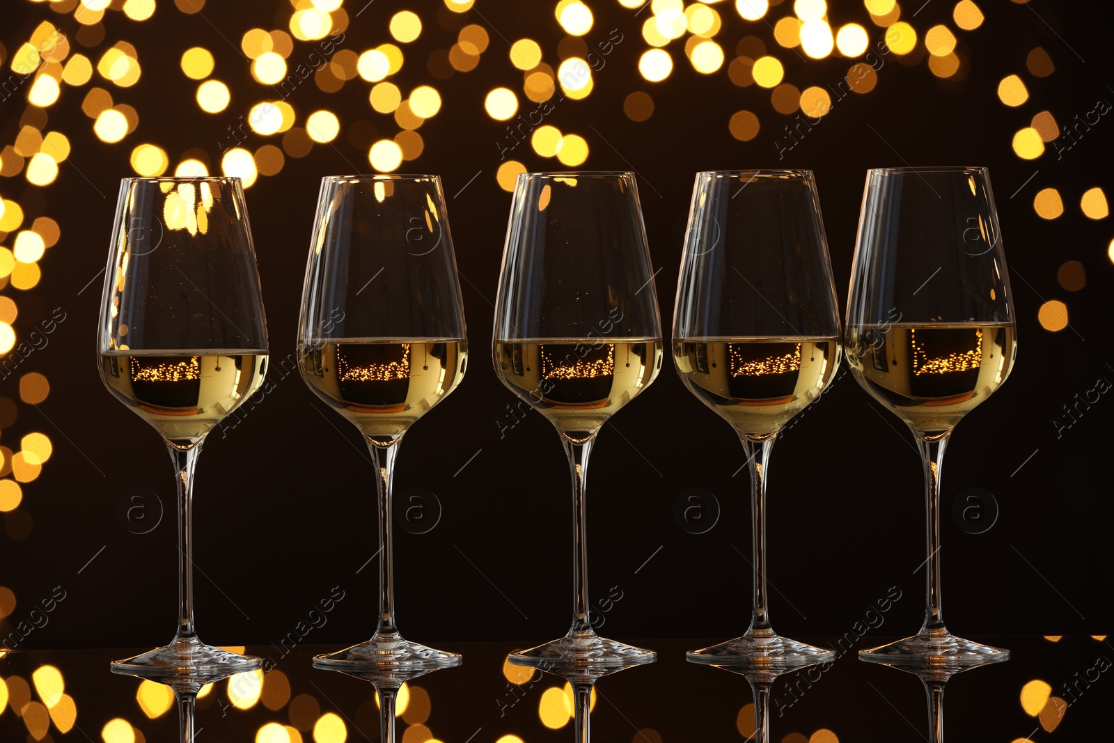 Photo of Tasty white wine in glasses on mirror surface against dark background with blurred lights. Bokeh effect