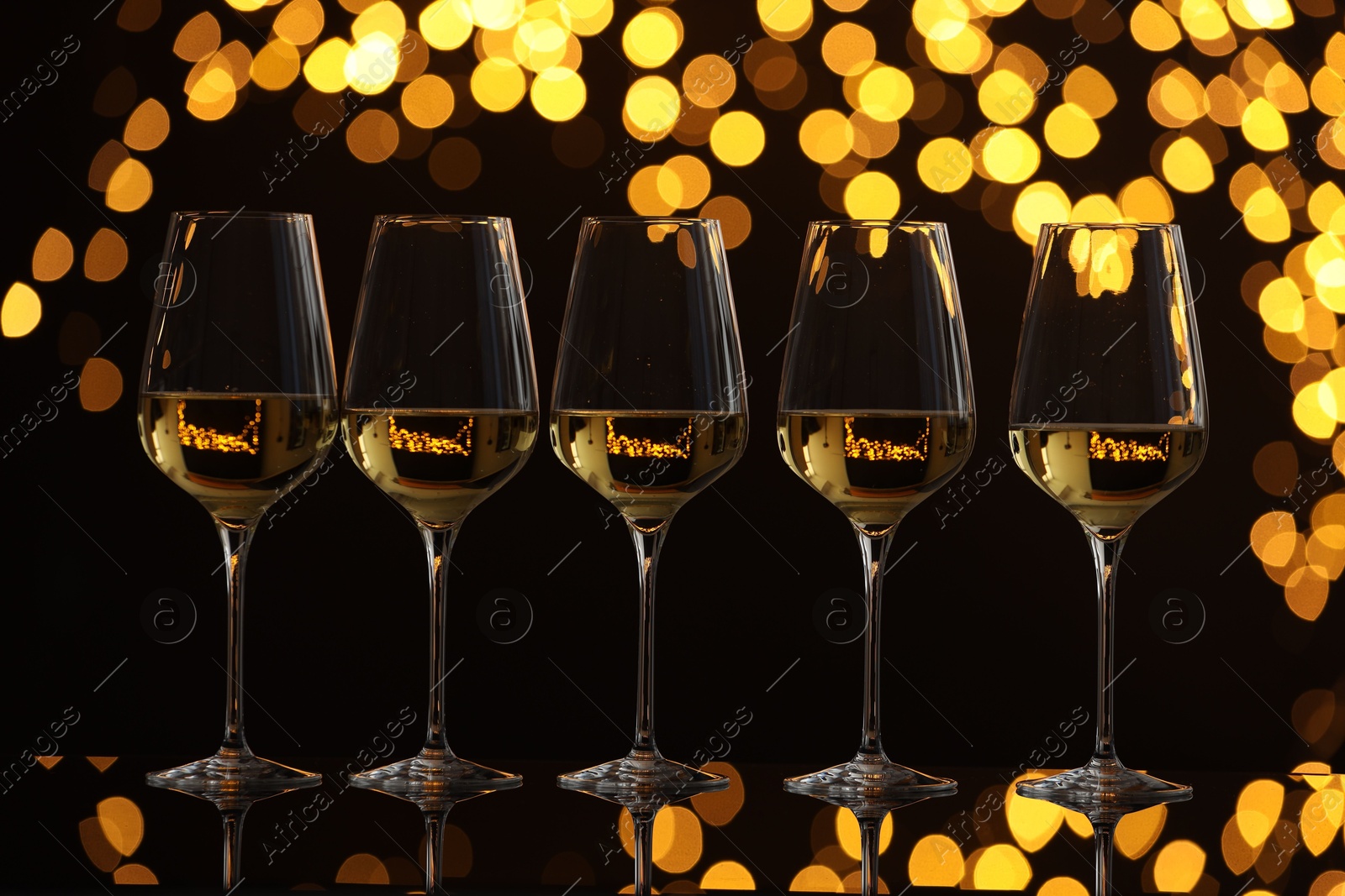 Photo of Tasty white wine in glasses on mirror surface against dark background with blurred lights. Bokeh effect