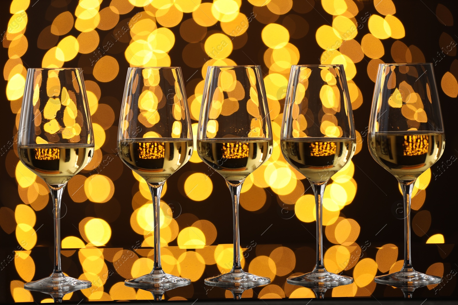 Photo of Tasty white wine in glasses on mirror surface against dark background with blurred lights. Bokeh effect