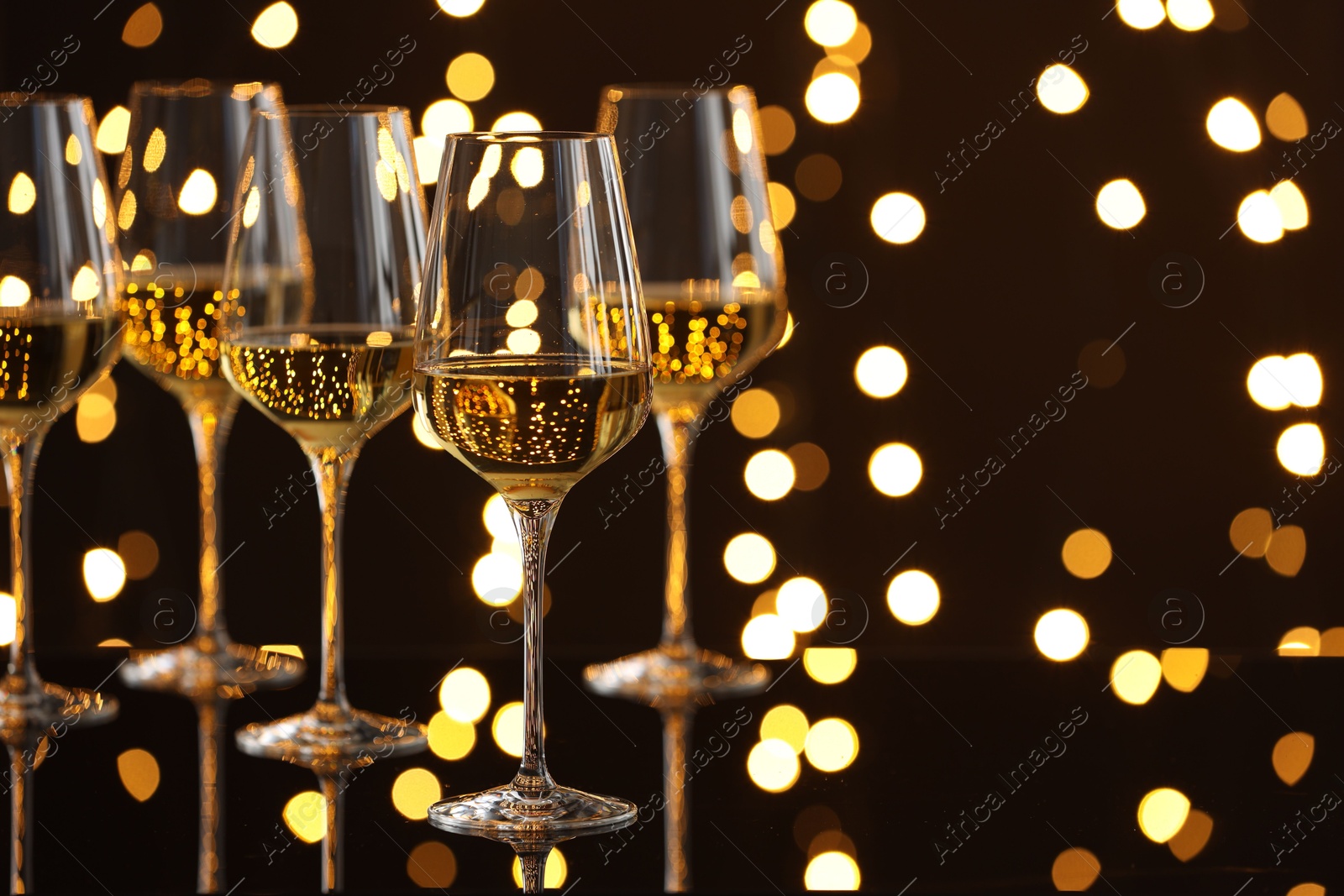 Photo of Tasty white wine in glasses on mirror surface against dark background with blurred lights, space for text. Bokeh effect