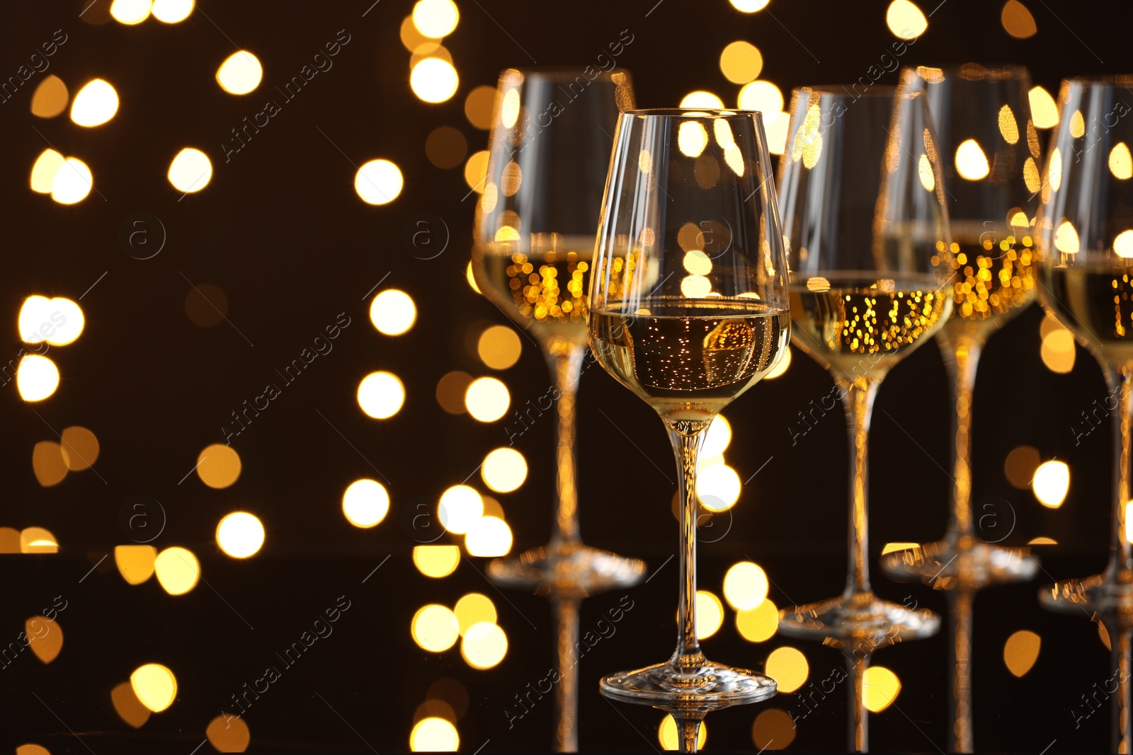 Photo of Tasty white wine in glasses on mirror surface against dark background with blurred lights, space for text. Bokeh effect