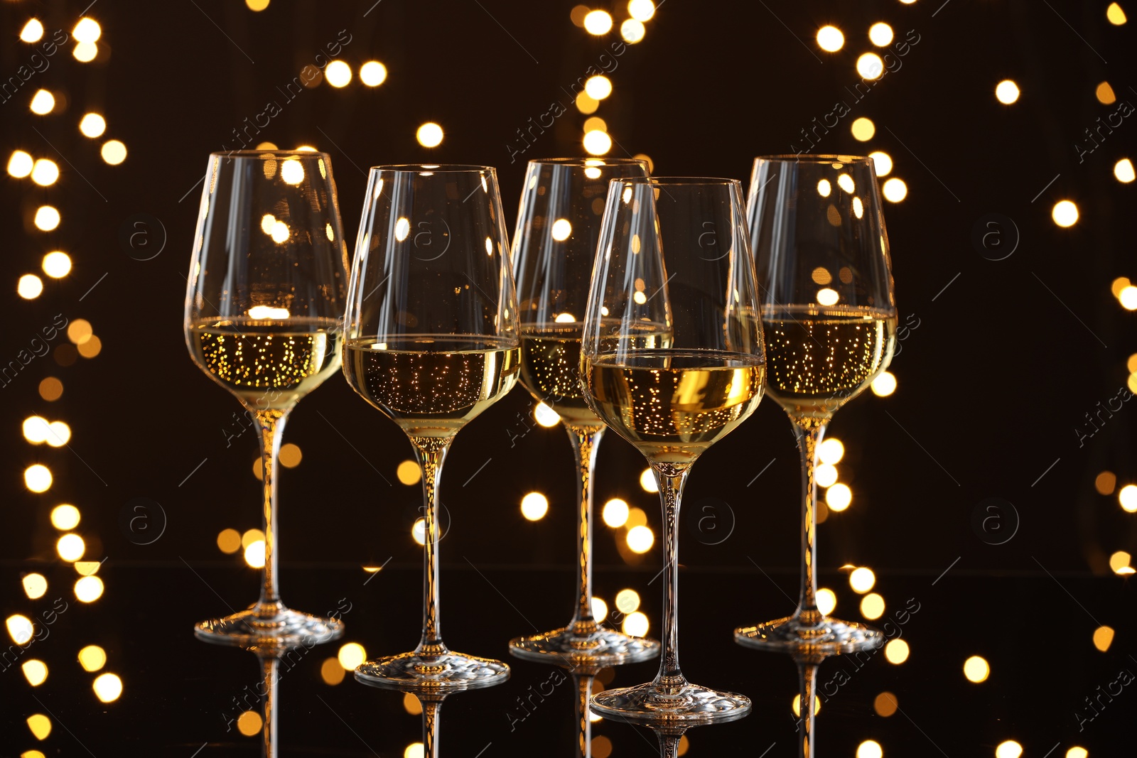 Photo of Tasty white wine in glasses on mirror surface against dark background with blurred lights. Bokeh effect