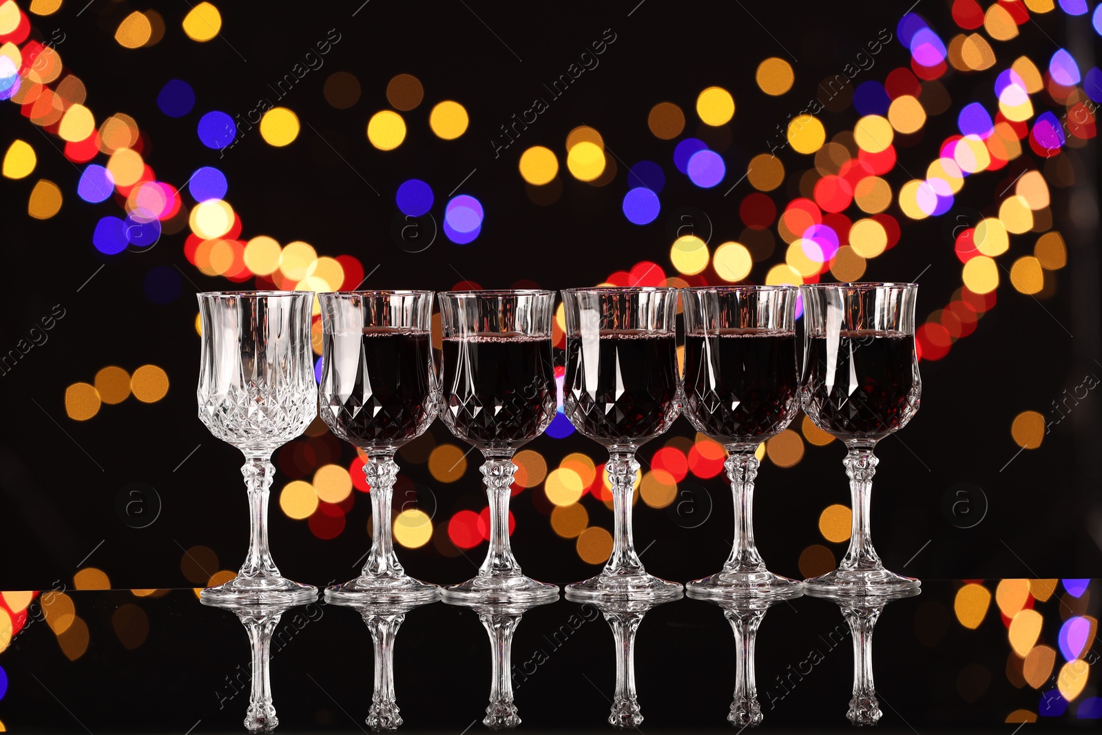 Photo of Tasty red wine in glasses and empty one on mirror surface against black background with blurred lights, bokeh effect