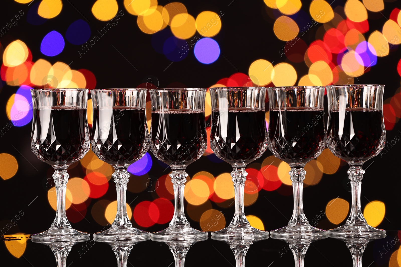 Photo of Tasty red wine in glasses on mirror surface against black background with blurred lights, bokeh effect