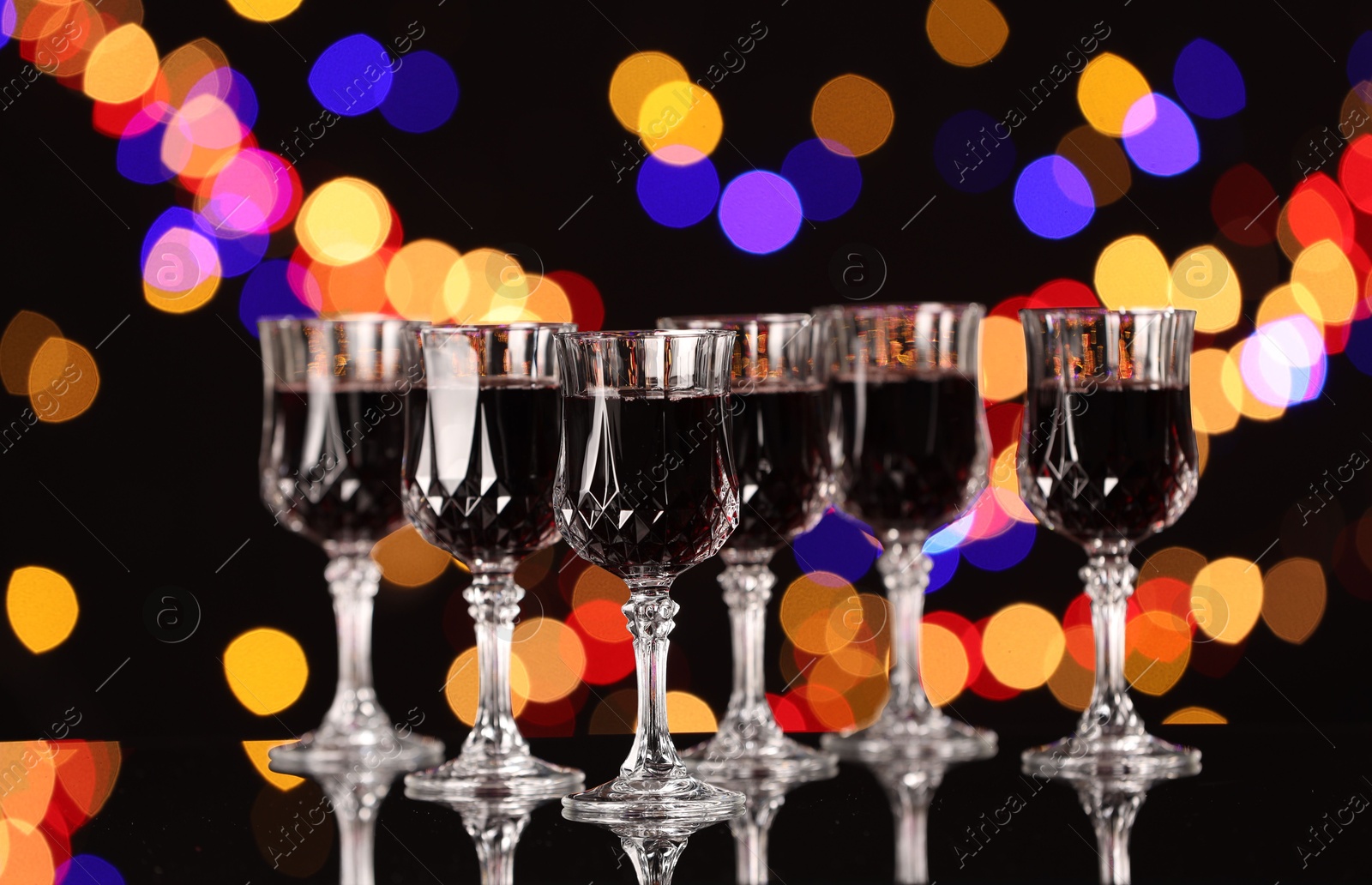 Photo of Tasty red wine in glasses on mirror surface against black background with blurred lights, bokeh effect