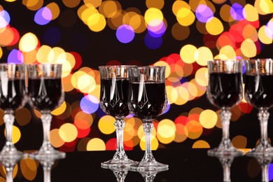 Photo of Tasty red wine in glasses on mirror surface against black background with blurred lights, bokeh effect