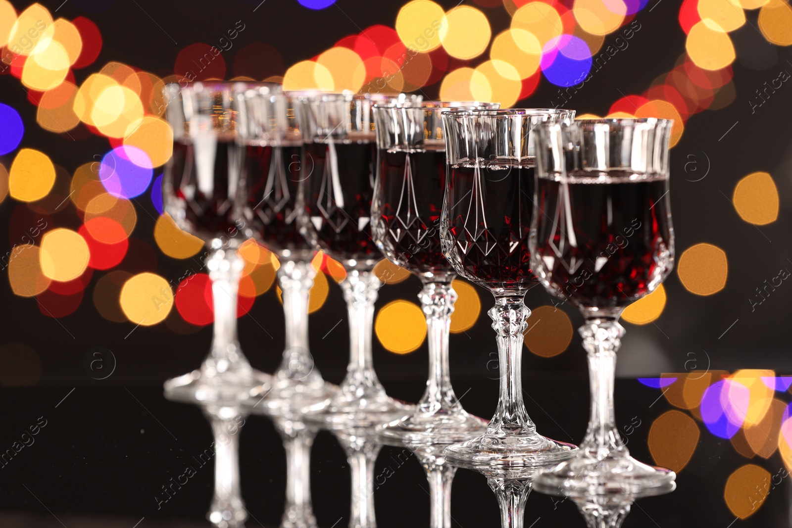 Photo of Tasty red wine in glasses on mirror surface against dark background with blurred lights, bokeh effect