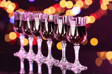 Photo of Tasty red wine in glasses on mirror surface against dark background with blurred lights, bokeh effect