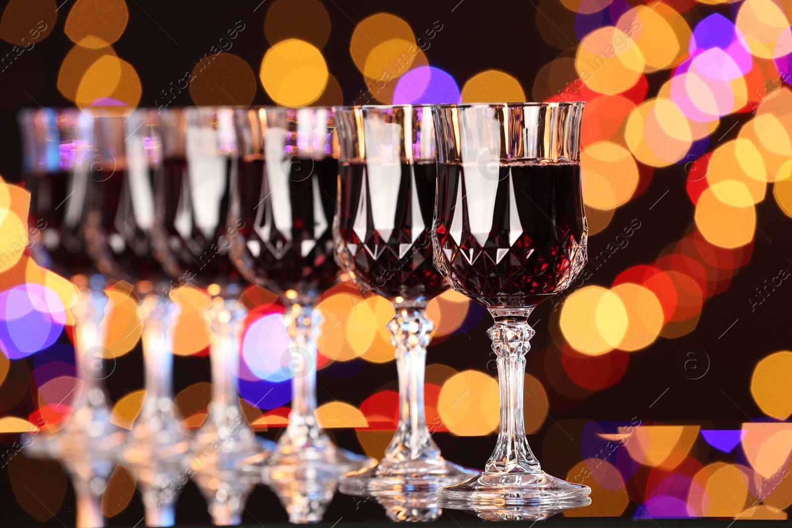 Photo of Tasty red wine in glasses on mirror surface against dark background with blurred lights, bokeh effect