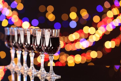 Photo of Tasty red wine in glasses on mirror surface against dark background with blurred lights, space for text. Bokeh effect