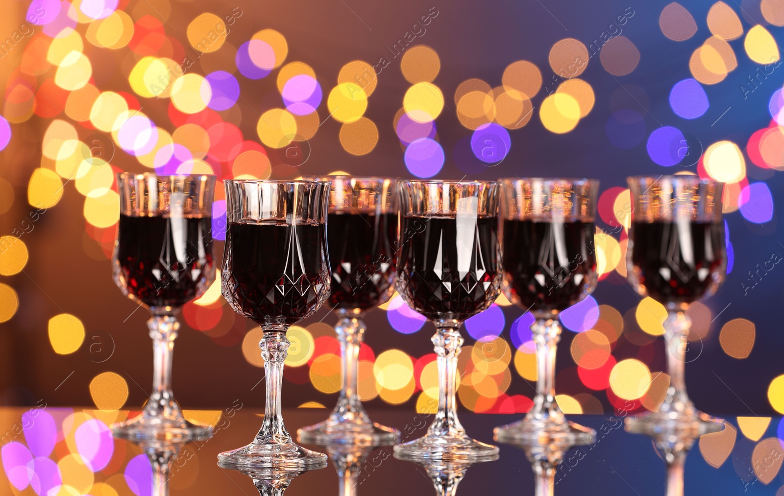 Photo of Tasty red wine in glasses on mirror surface against dark background with blurred lights, bokeh effect