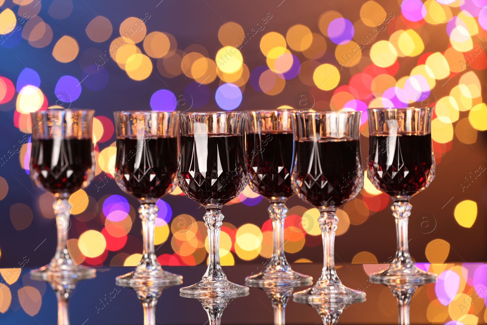 Photo of Tasty red wine in glasses on mirror surface against dark background with blurred lights, bokeh effect