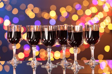 Photo of Tasty red wine in glasses on mirror surface against dark background with blurred lights, bokeh effect