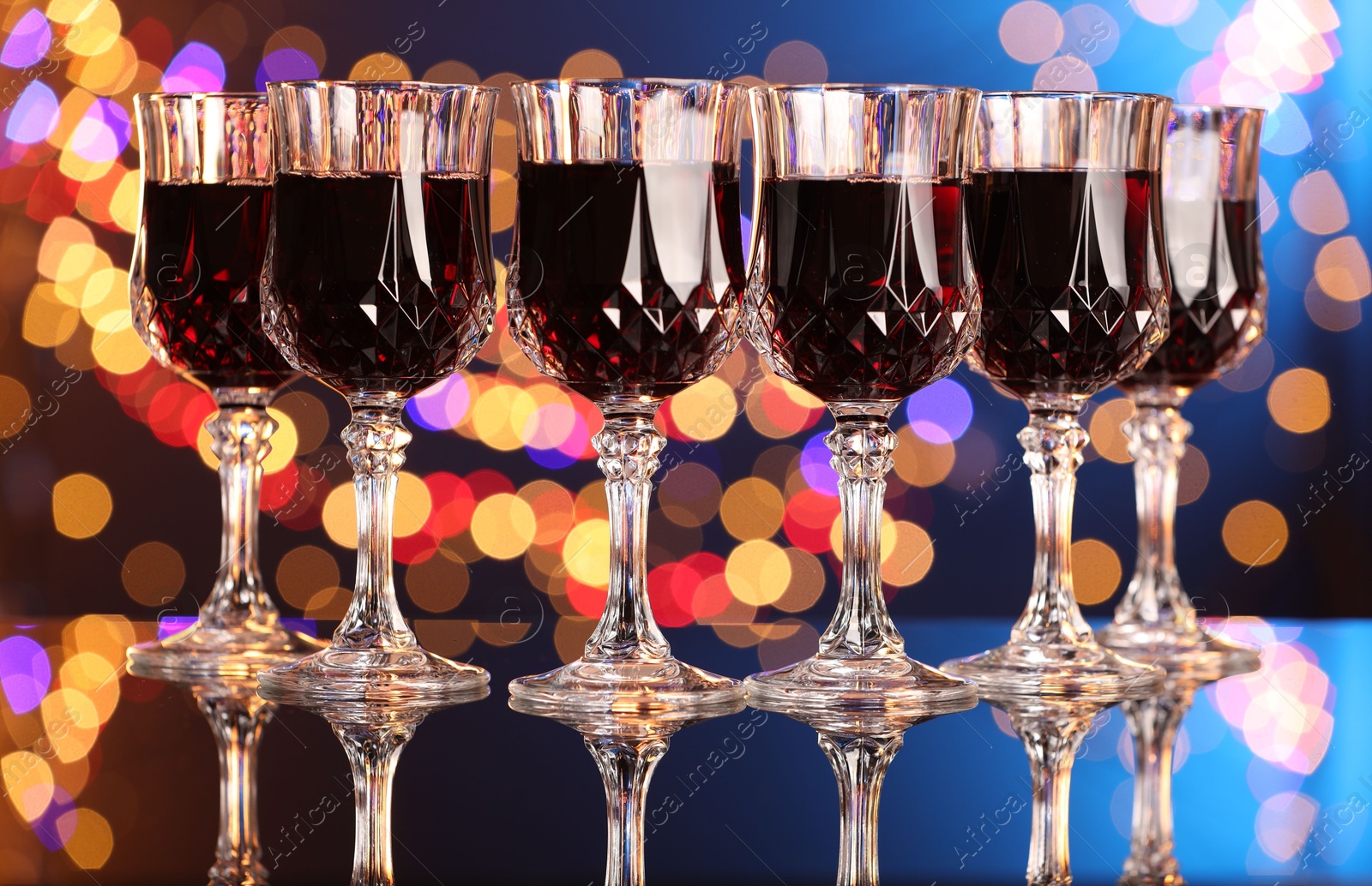 Photo of Tasty red wine in glasses on mirror surface against color background with blurred lights, bokeh effect