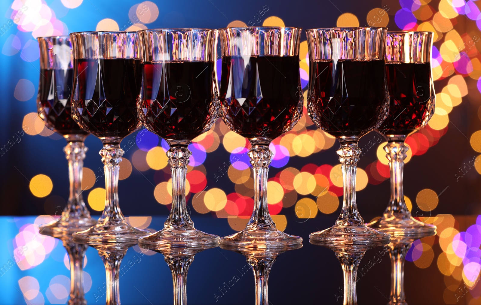 Photo of Tasty red wine in glasses on mirror surface against color background with blurred lights, bokeh effect