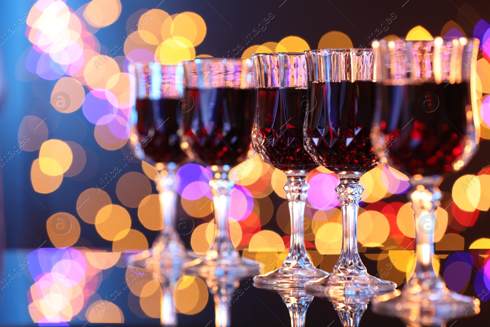 Photo of Tasty red wine in glasses on mirror surface against dark background with blurred lights, bokeh effect