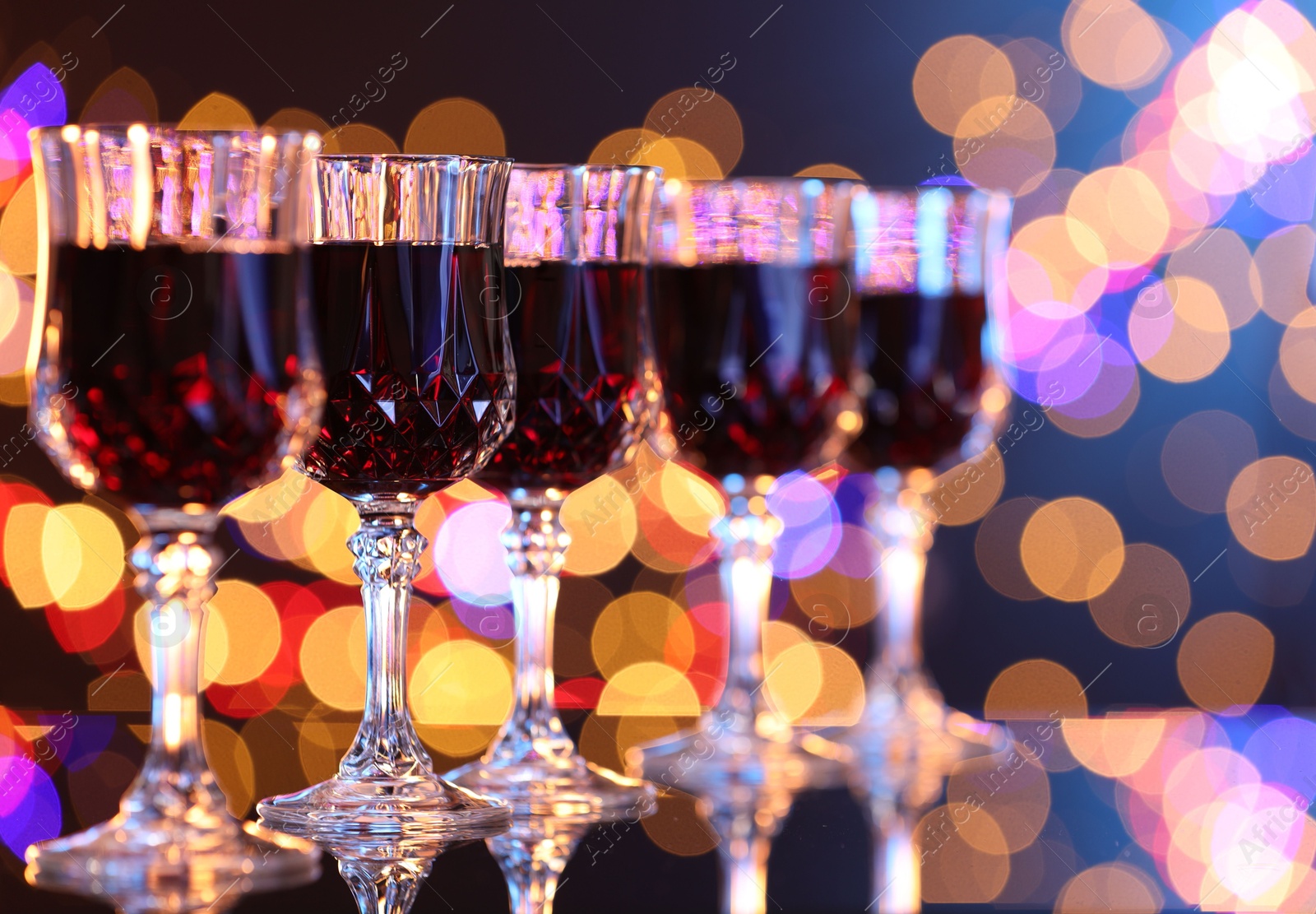 Photo of Tasty red wine in glasses on mirror surface against dark background with blurred lights, bokeh effect