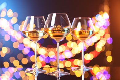 Photo of Tasty white wine in glasses on mirror surface against dark background with blurred lights, bokeh effect