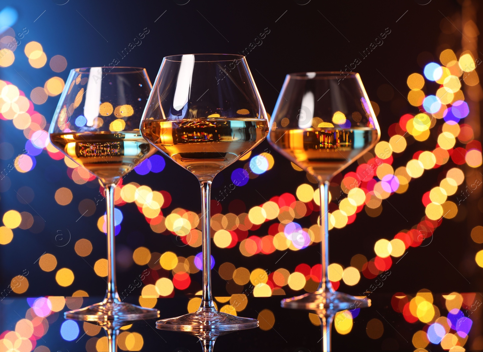 Photo of Tasty white wine in glasses on mirror surface against dark background with blurred lights, bokeh effect