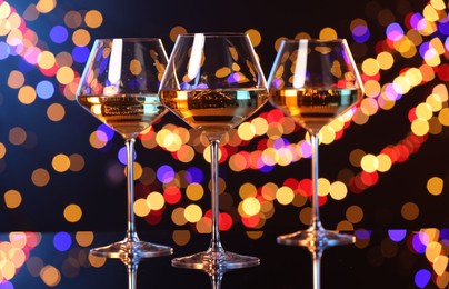 Photo of Tasty white wine in glasses on mirror surface against dark background with blurred lights, bokeh effect