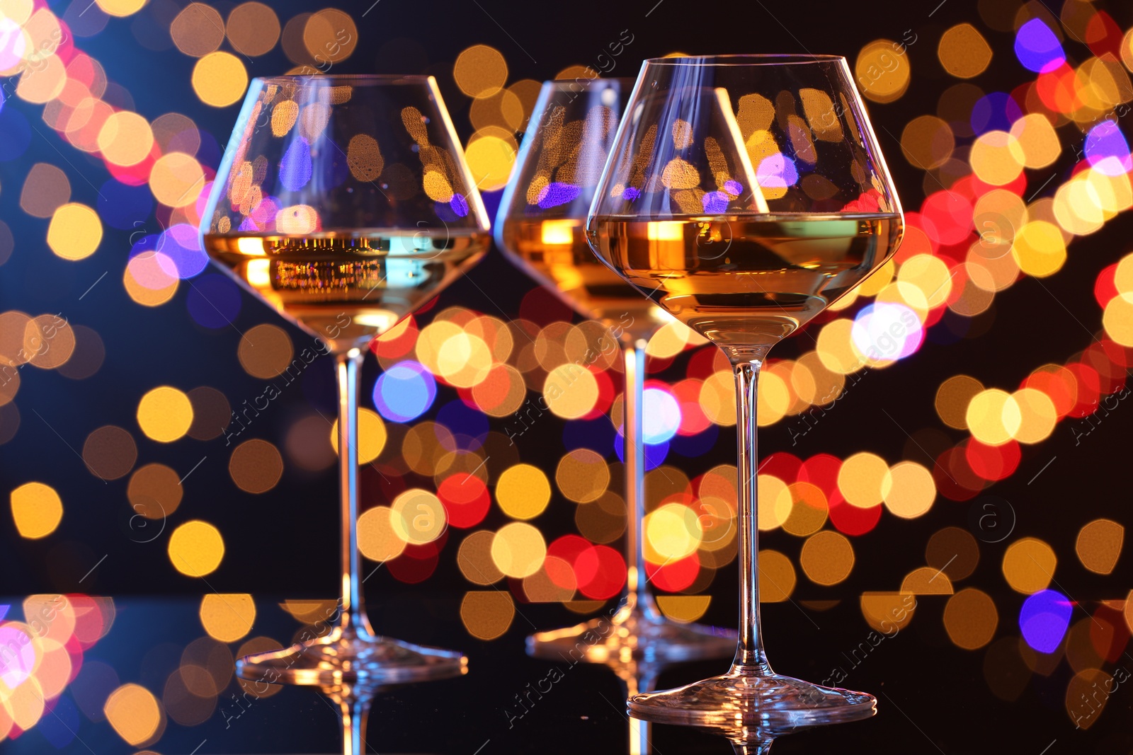 Photo of Tasty white wine in glasses on mirror surface against dark background with blurred lights, bokeh effect