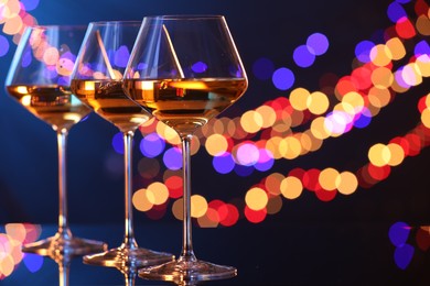 Photo of Tasty white wine in glasses on mirror surface against dark blue background with blurred lights, space for text. Bokeh effect