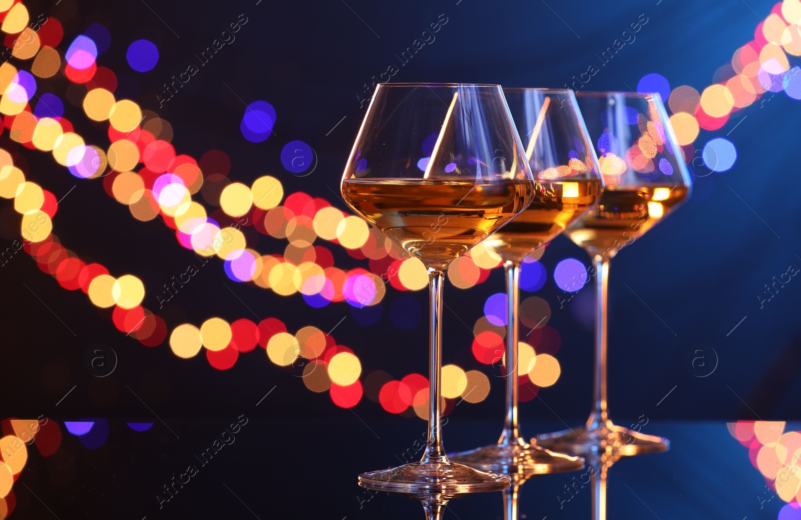 Photo of Tasty white wine in glasses on mirror surface against dark blue background with blurred lights, space for text. Bokeh effect