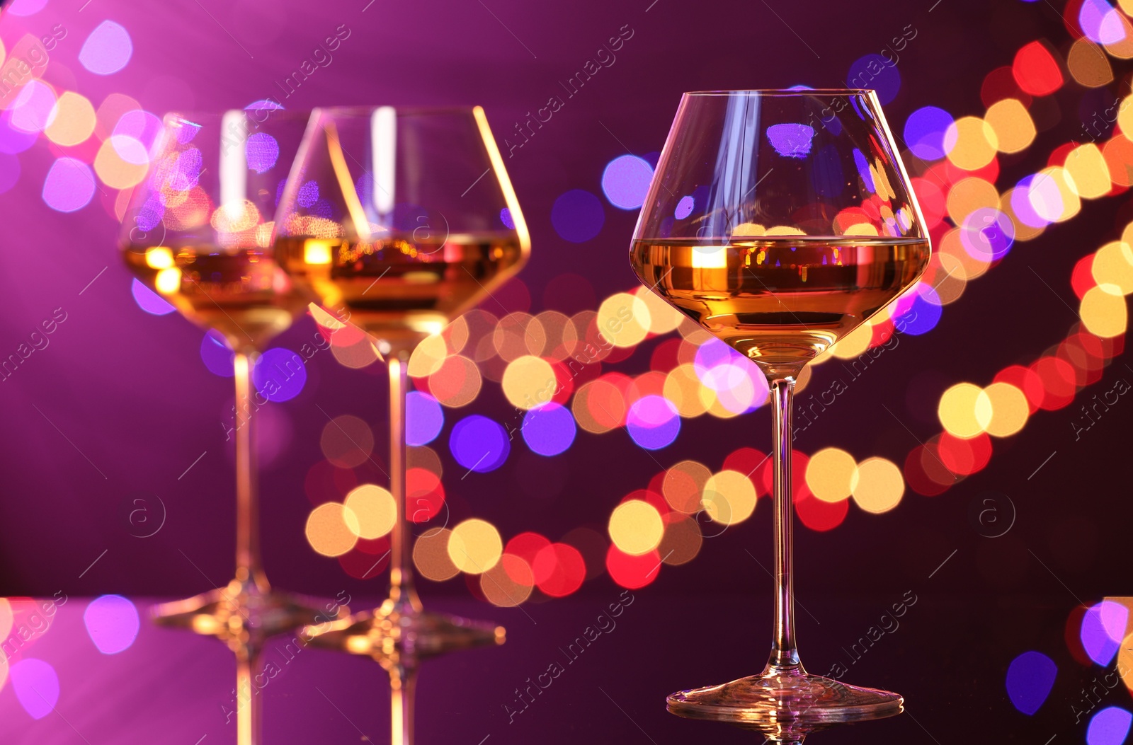 Photo of Tasty white wine in glasses on mirror surface against purple background with blurred lights, bokeh effect