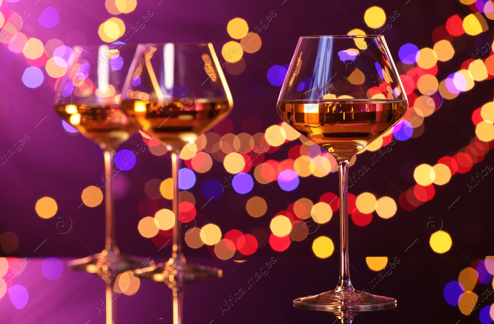 Photo of Tasty white wine in glasses on mirror surface against purple background with blurred lights, bokeh effect