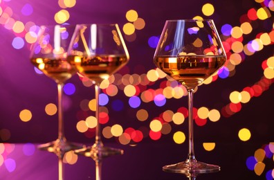 Photo of Tasty white wine in glasses on mirror surface against purple background with blurred lights, bokeh effect