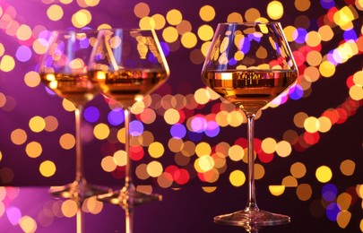 Photo of Tasty white wine in glasses on mirror surface against purple background with blurred lights, bokeh effect