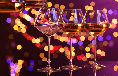 Photo of Pouring white wine into glass against purple background with blurred lights, closeup. Bokeh effect